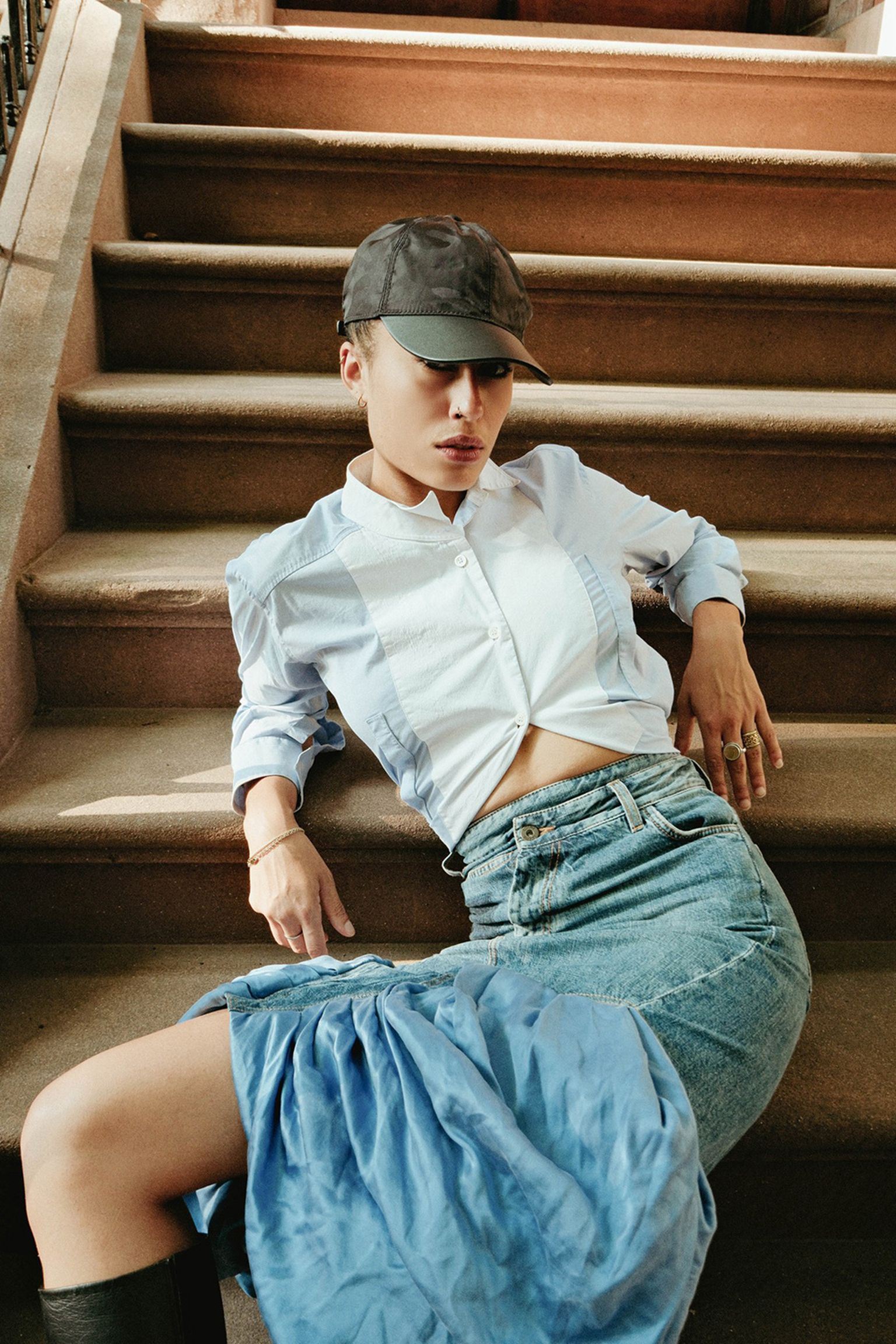 Person in a black cap and white shirt, leaning back on stairs, wearing a denim skirt and blue fabric draped over the knee