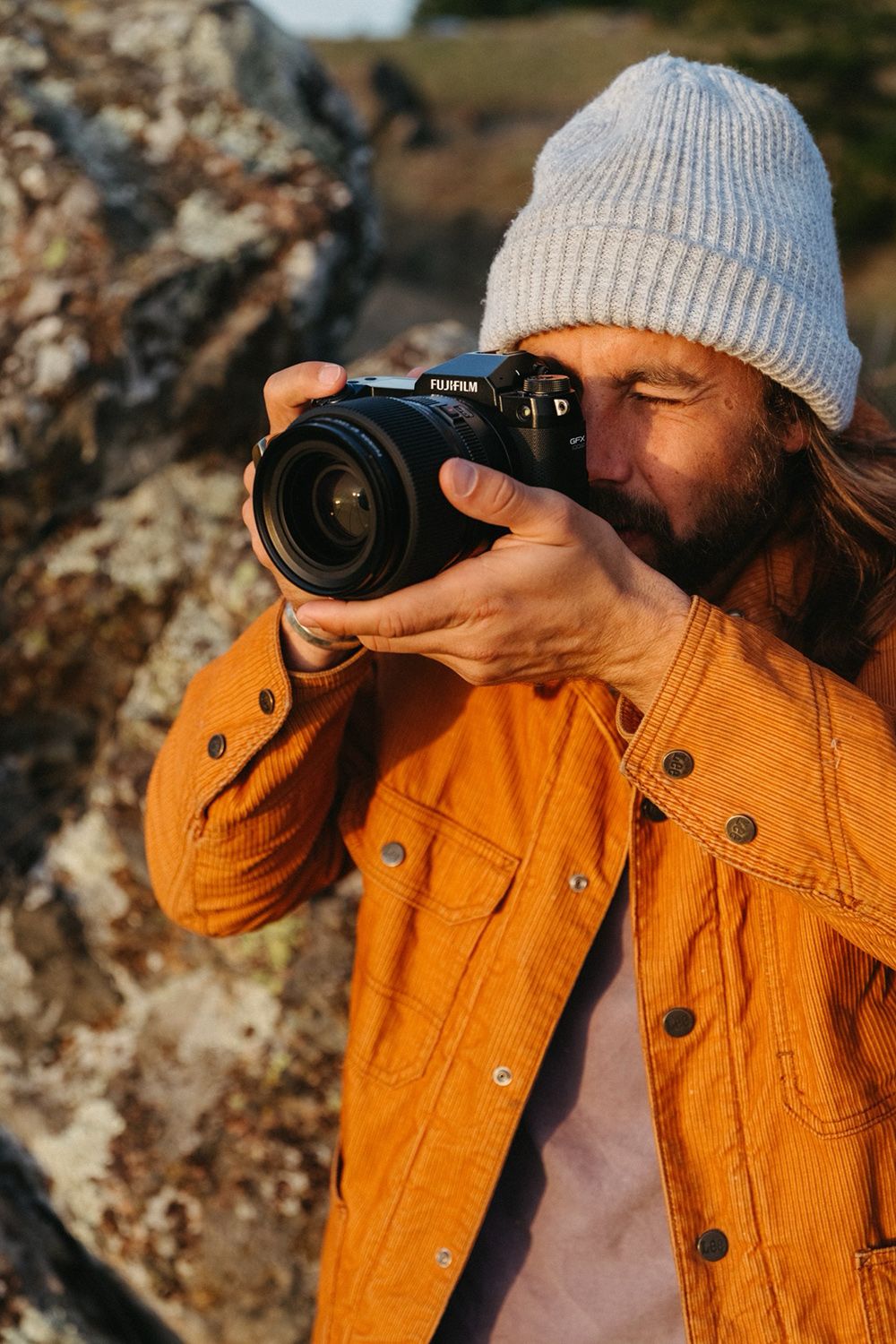 A person in a yellow jacket and gray beanie makes a photo with a Fujifilm camera outdoors, near rocks