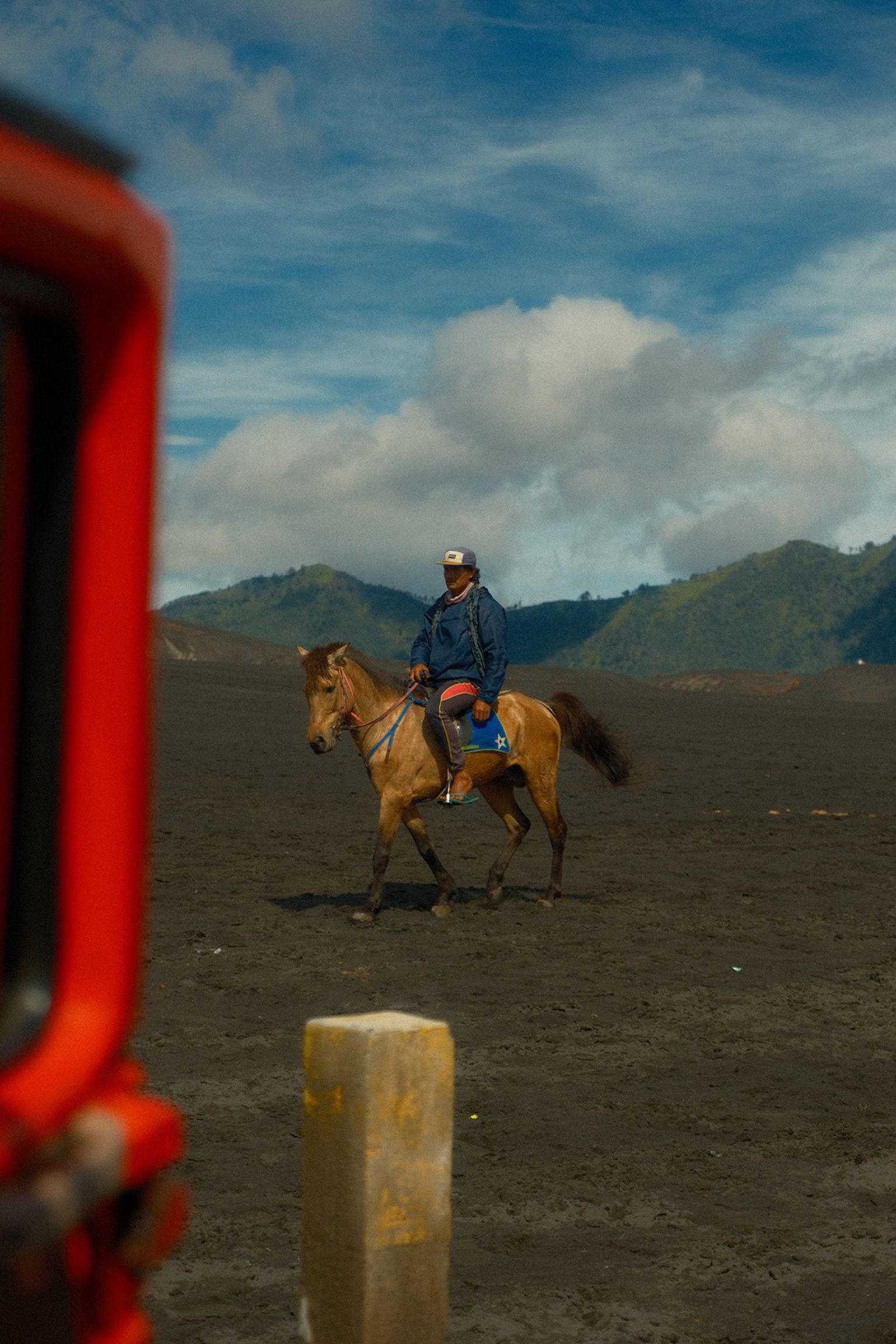A person rides a horse across a barren, dark sandy plain with mountains and a cloudy sky in the background, and a red vehicle partially visible