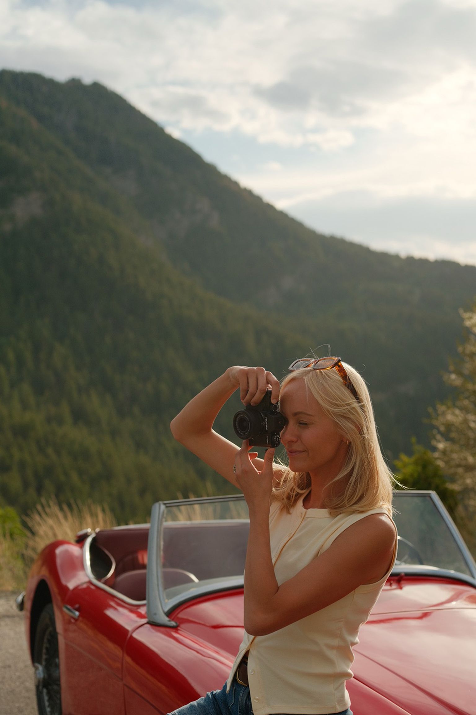 Woman photographing with a camera beside a red convertible in a mountainous landscape