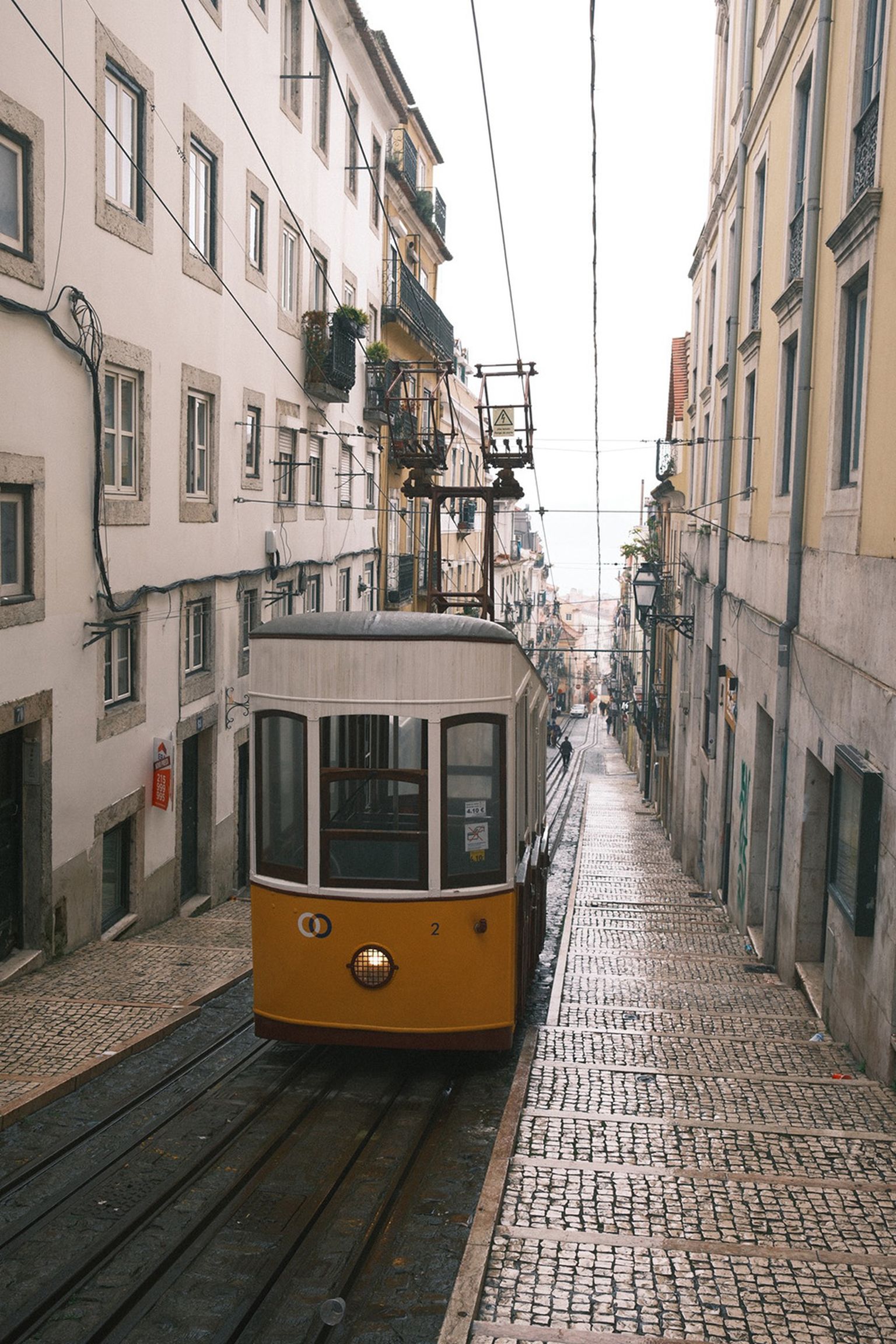 Yellow tram on a steep, cobblestone street lined with historic buildings, with power lines overhead