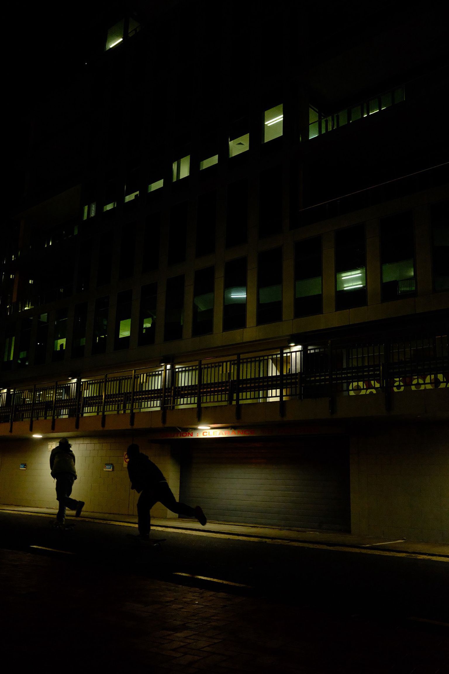 Two people run on a dimly lit street at night, with a tall building in the background illuminated by internal lights