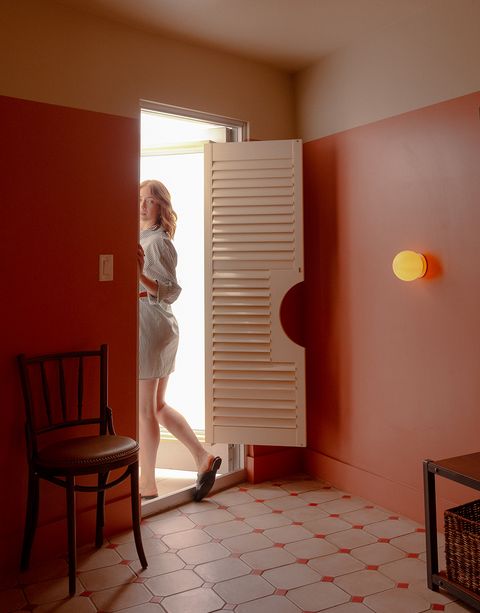 A woman in a striped dress stands in a doorway with a half-open shutter door in a room with terracotta walls and tiled floor