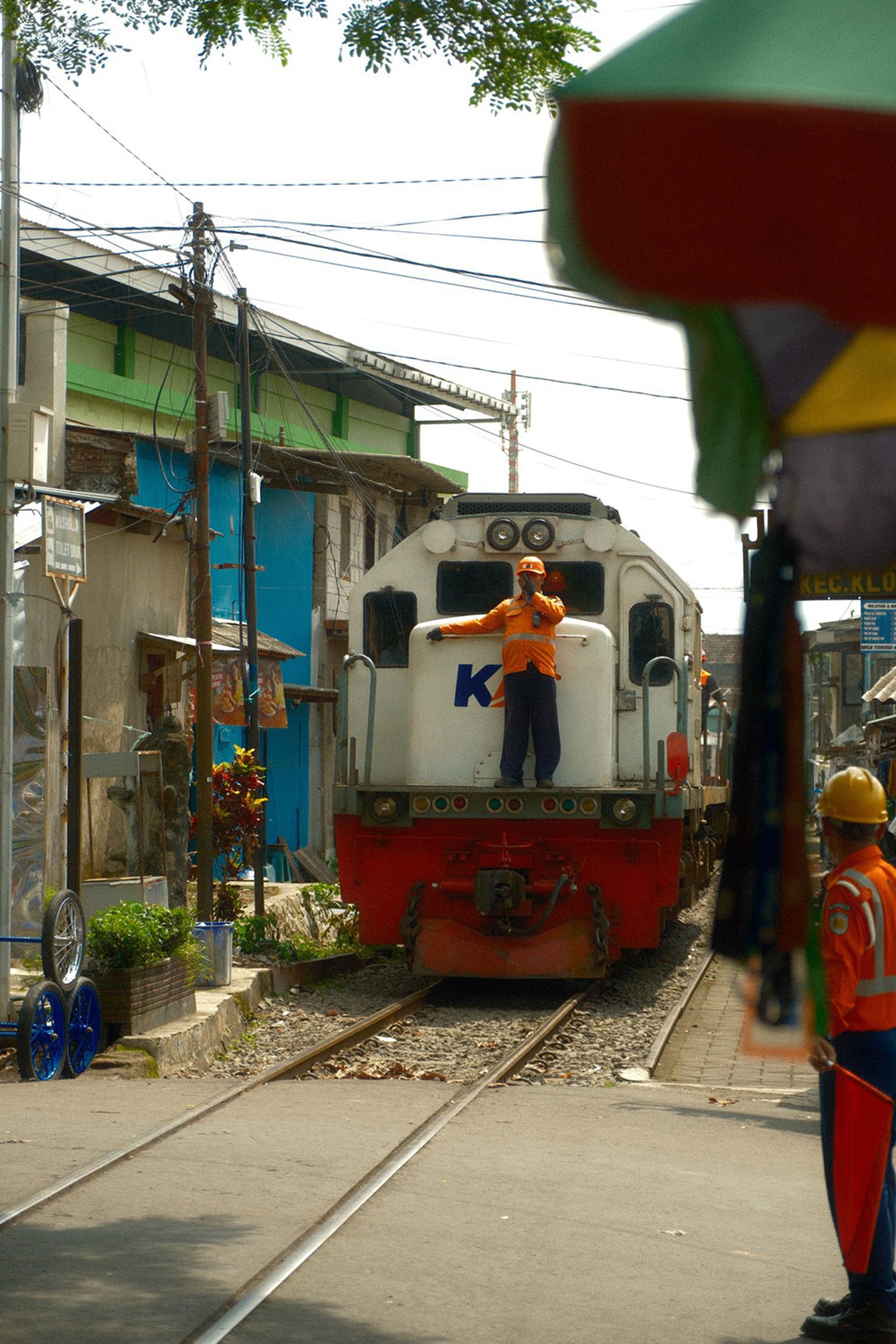 A man in an orange vest stands on the front of a stationary train, with buildings and other workers visible nearby