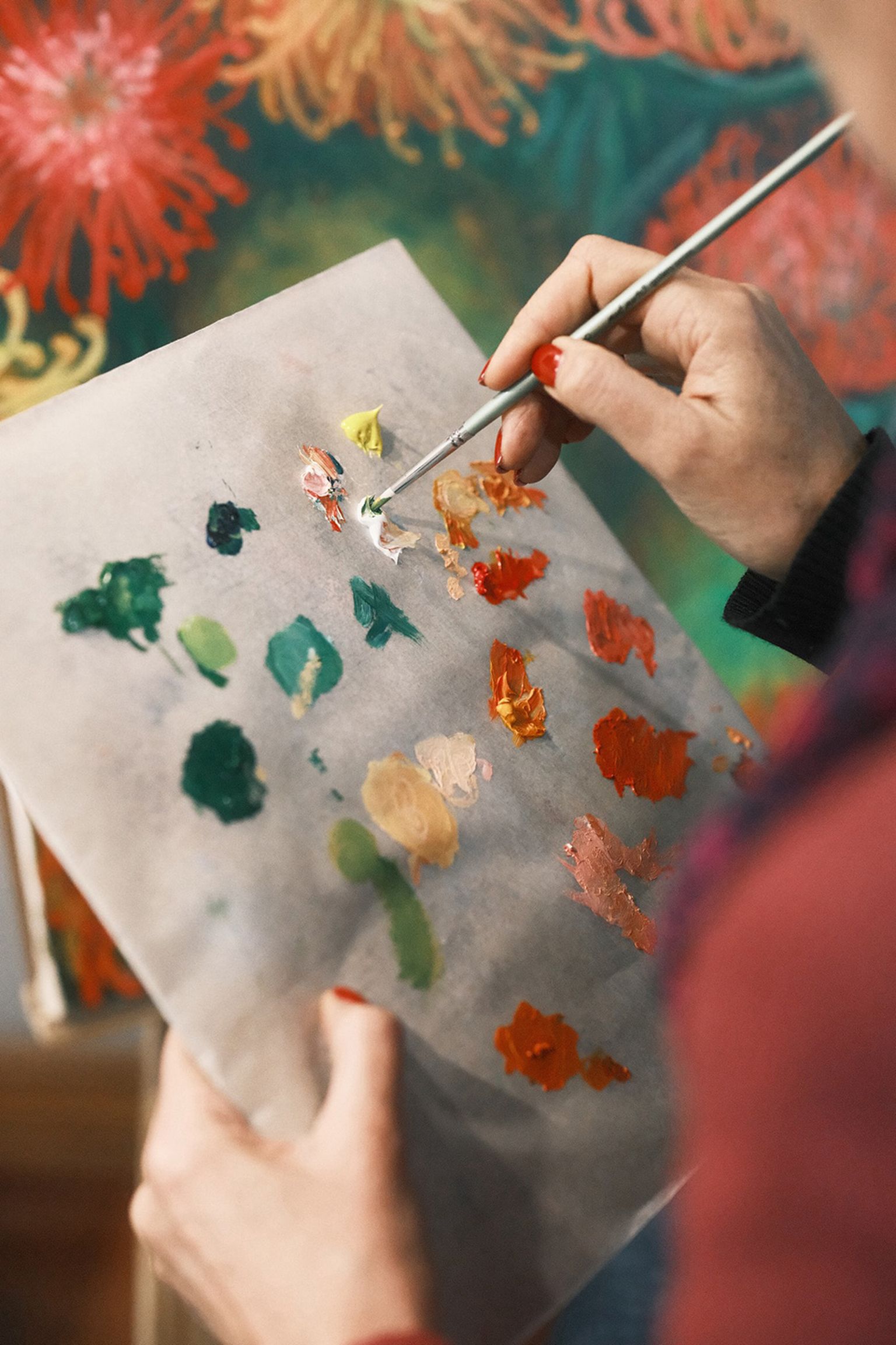 Close-up of an artist's hand holding a brush, mixing vibrant paints on a palette, with a colorful painting in the background