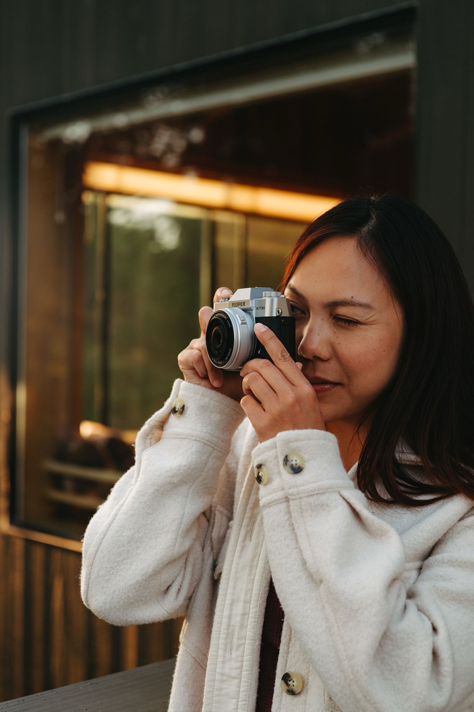A woman in a light coat is creating a photo with a camera near a reflective window