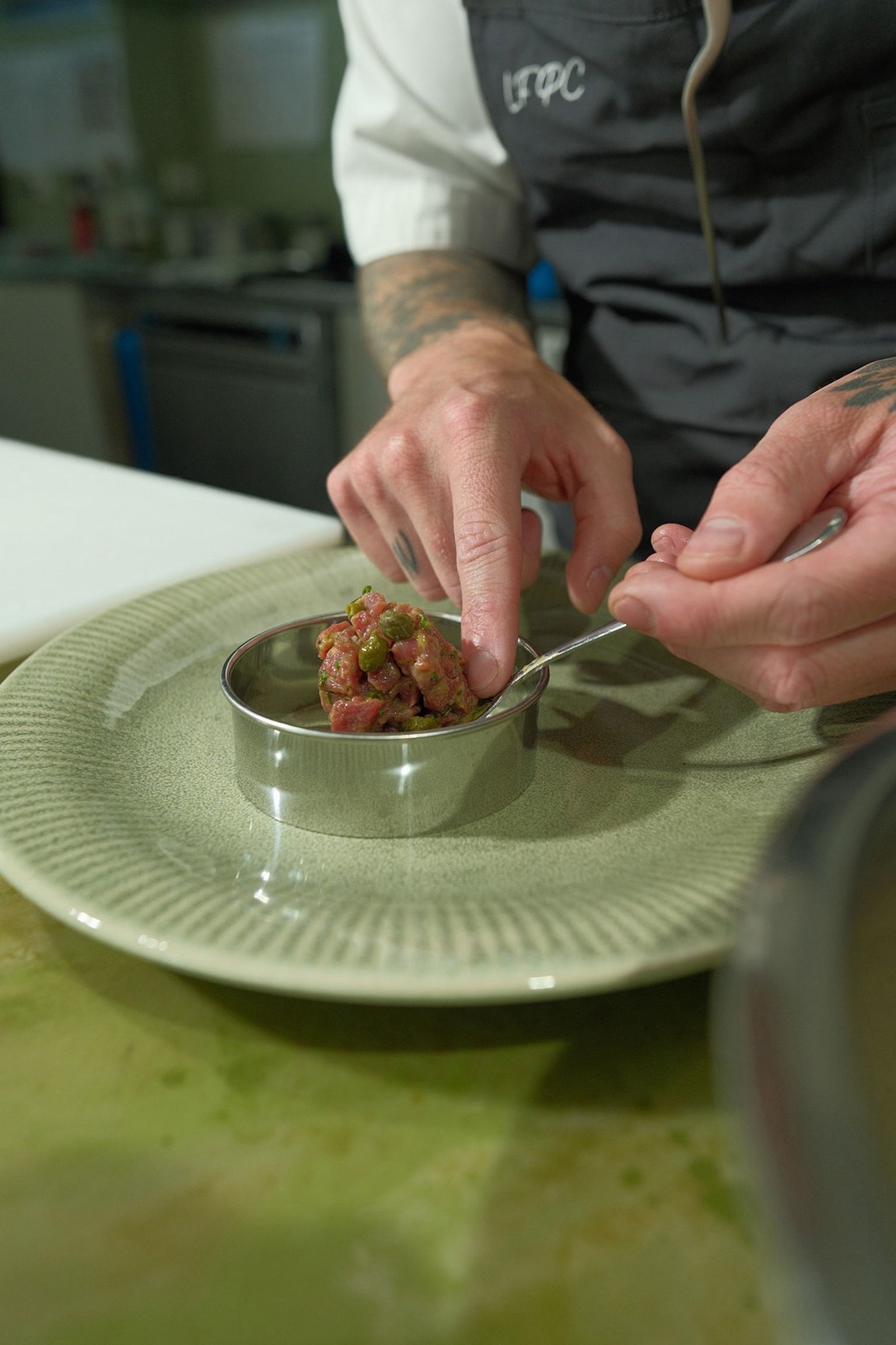 Chef preparing tartare in a metal ring on a green plate, using a spoon to shape the ingredients