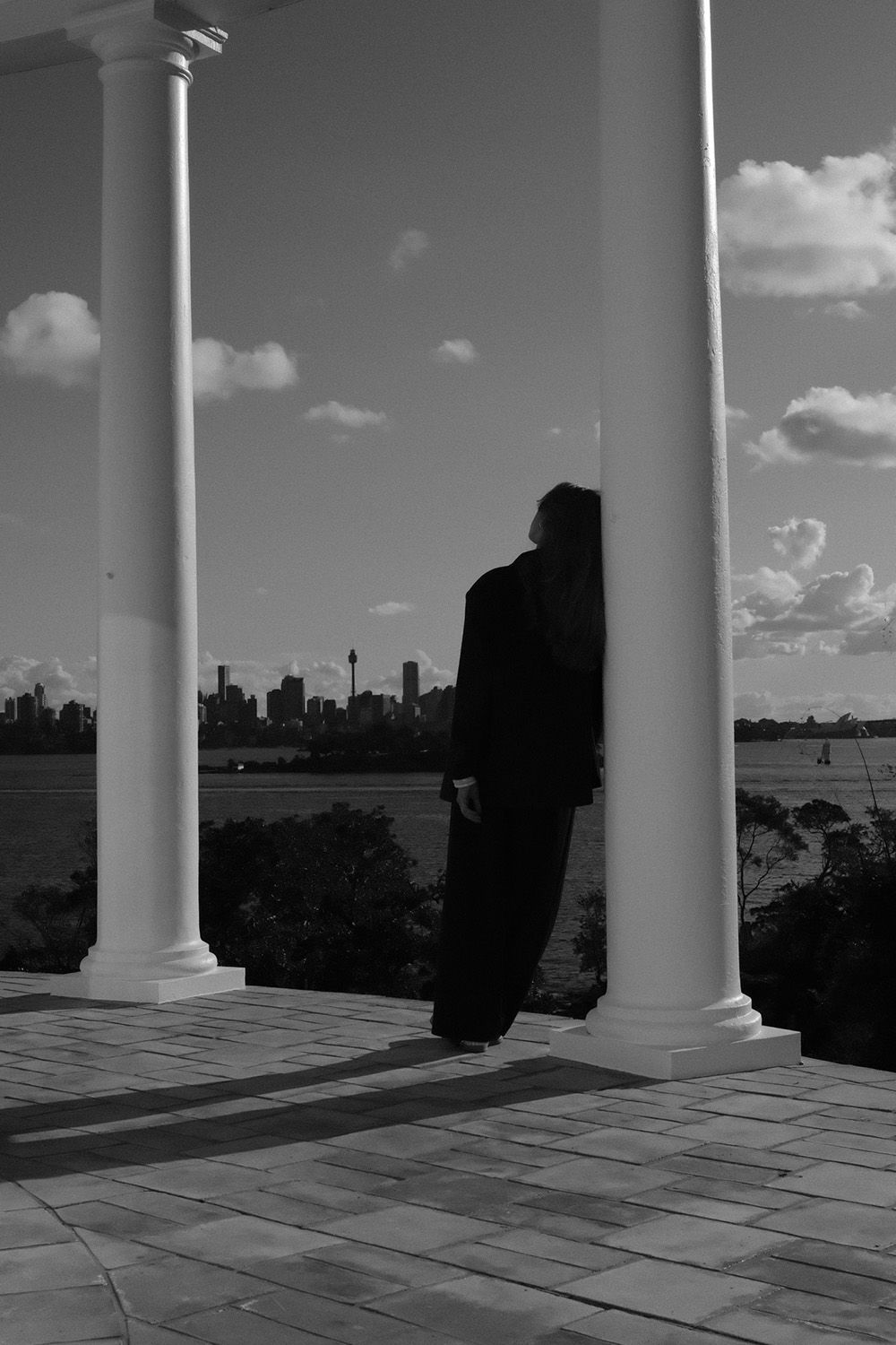 Person in dark clothing leans against a column on a terrace, overlooking a city skyline and water under a cloudy sky