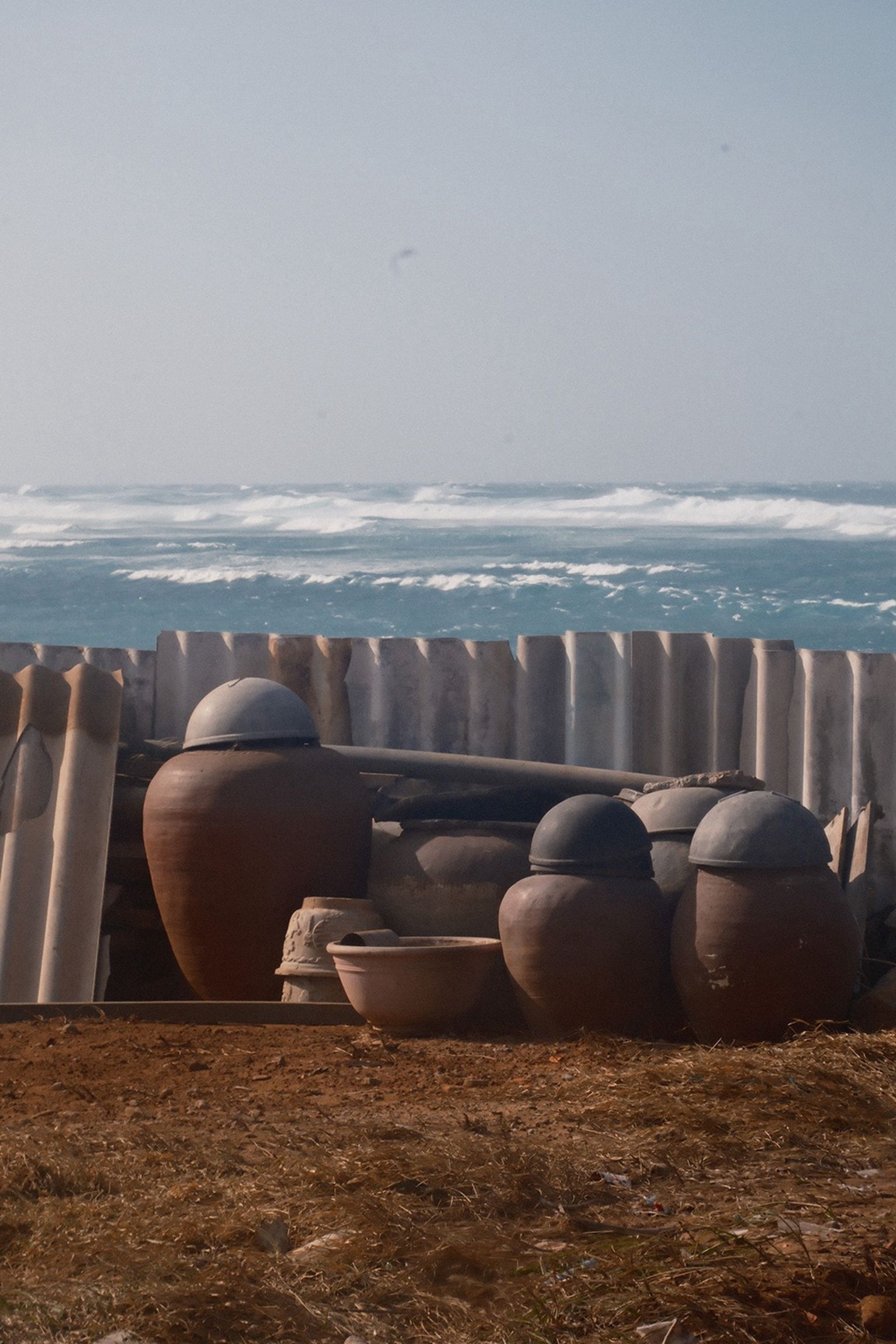Large clay pots and assorted pottery against a backdrop of a wooden fence, with ocean waves and a clear sky in the background