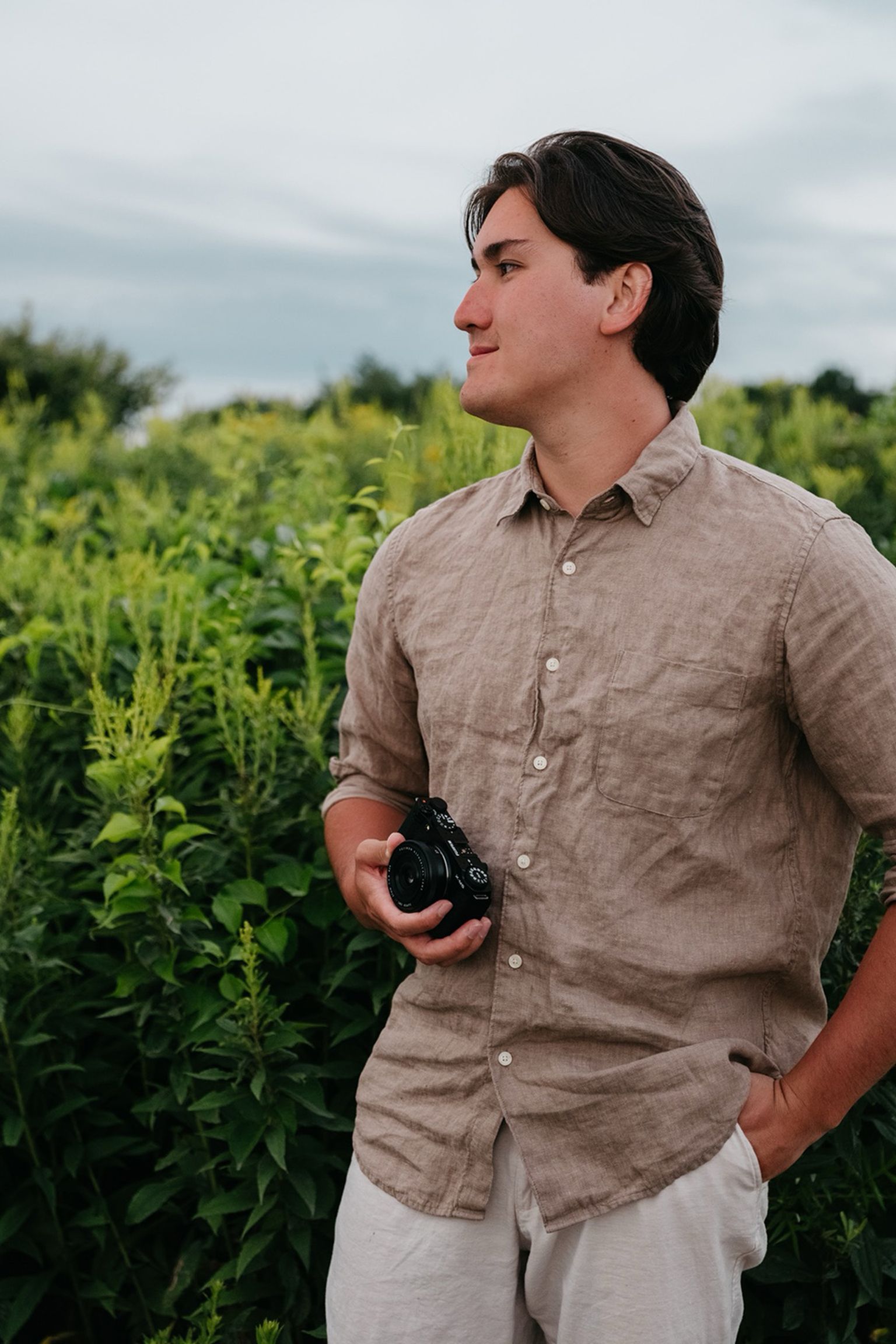 A man in a beige shirt holds a camera and looks to the side while standing in a lush green field under a cloudy sky