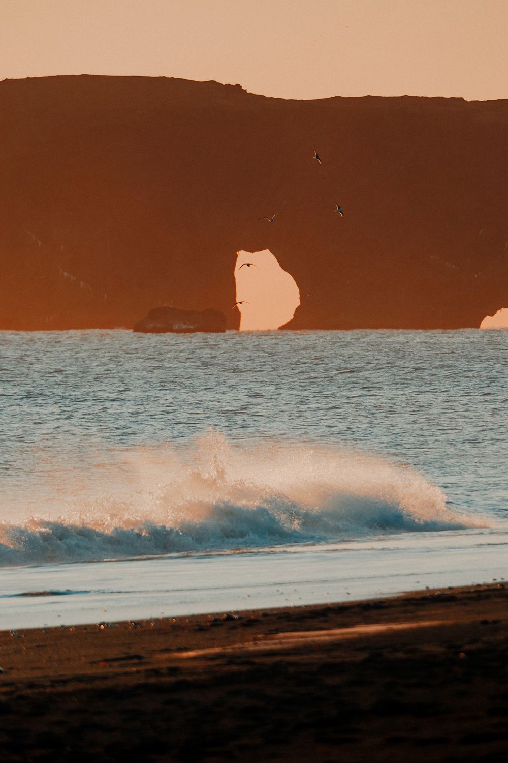 Waves crash on a dark sandy beach with a rock arch in the background under a clear sky, silhouetted by the setting sun