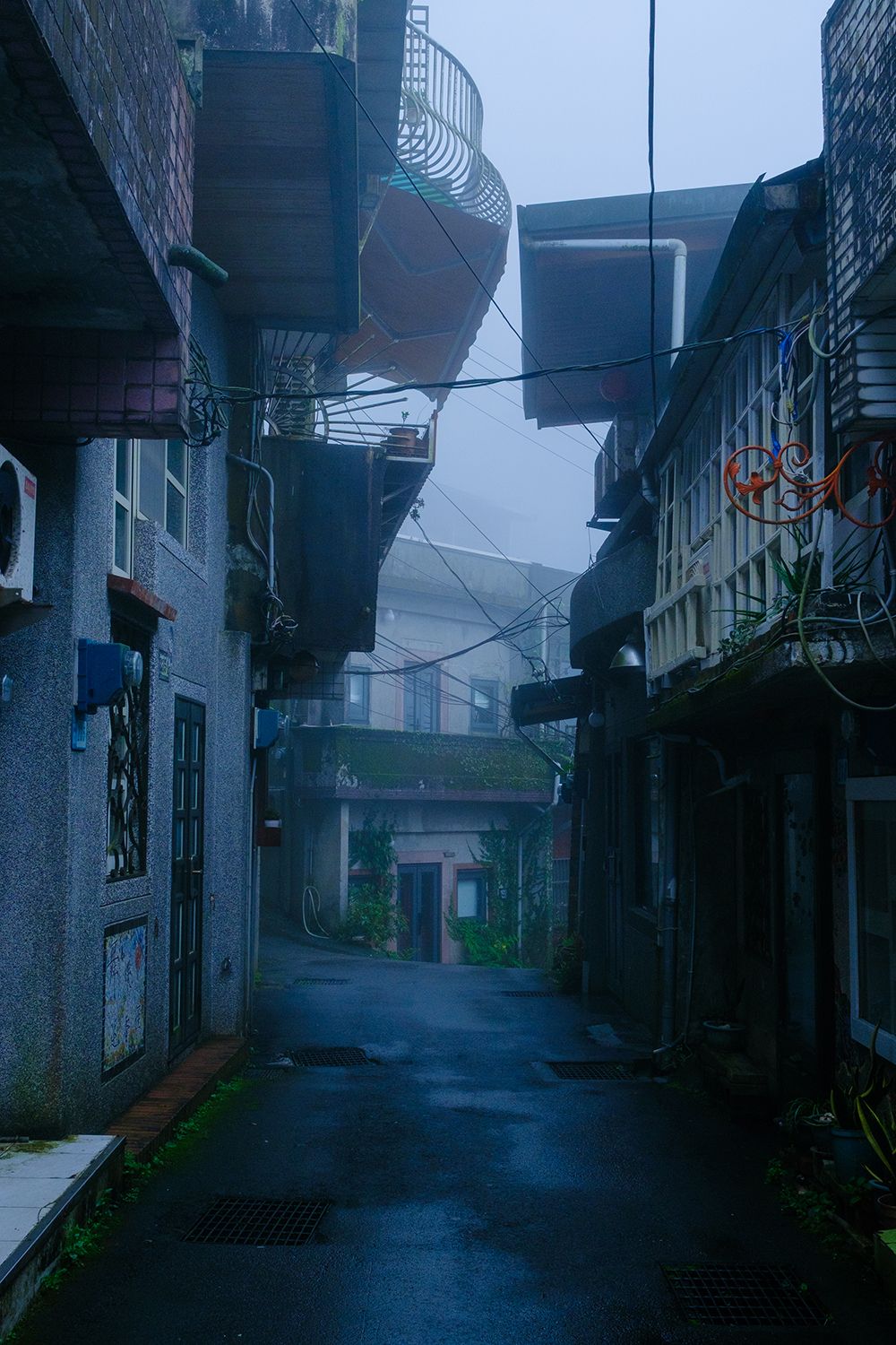 Narrow, misty alleyway between weathered buildings with overhanging balconies and tangled wires, creating a mysterious urban scene