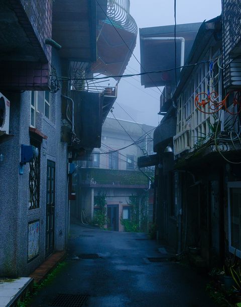 Narrow, misty alleyway between weathered buildings with overhanging balconies and tangled wires, creating a mysterious urban scene