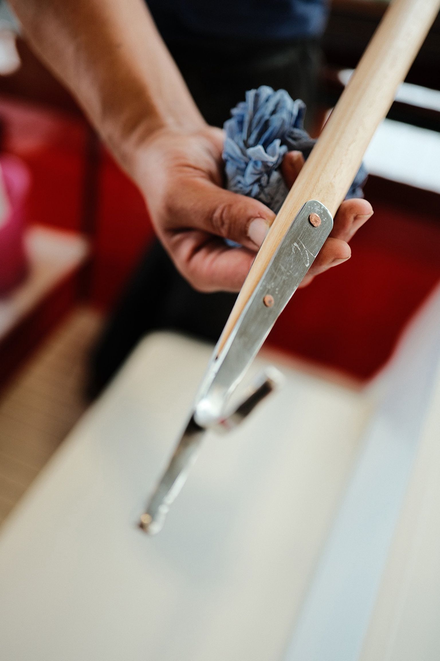 A person holding a blue cloth and a tool with a wooden handle and metal end, against a blurred red background