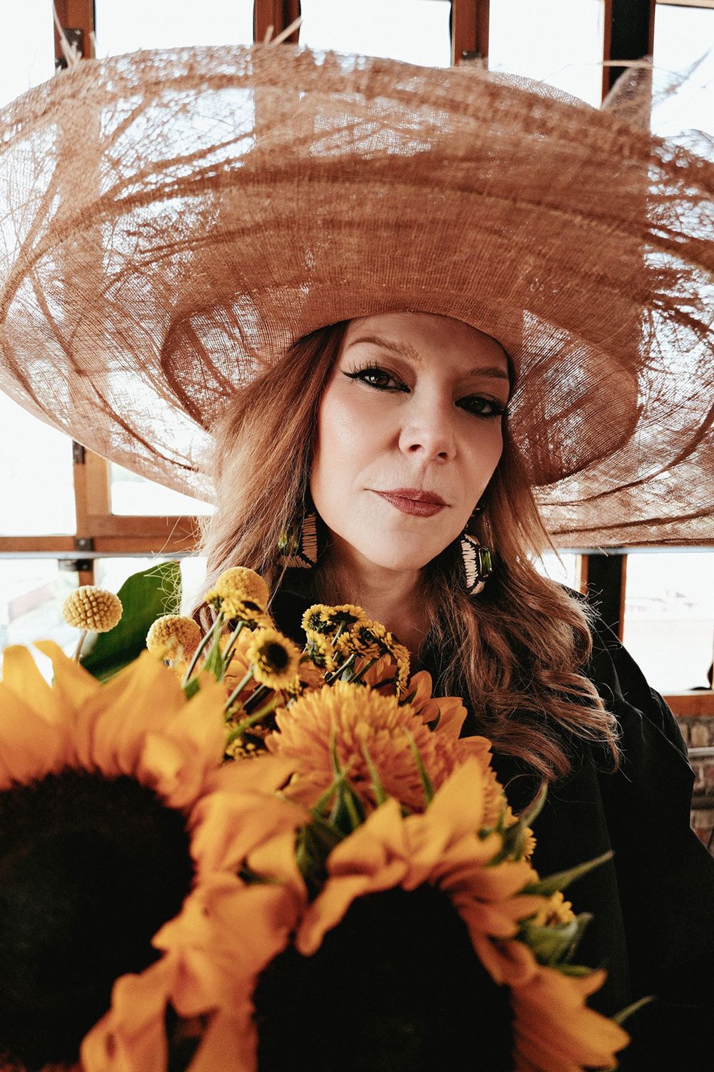 A woman wearing a large, woven hat holds a bouquet of vibrant sunflowers and yellow flowers, staring intently at the camera