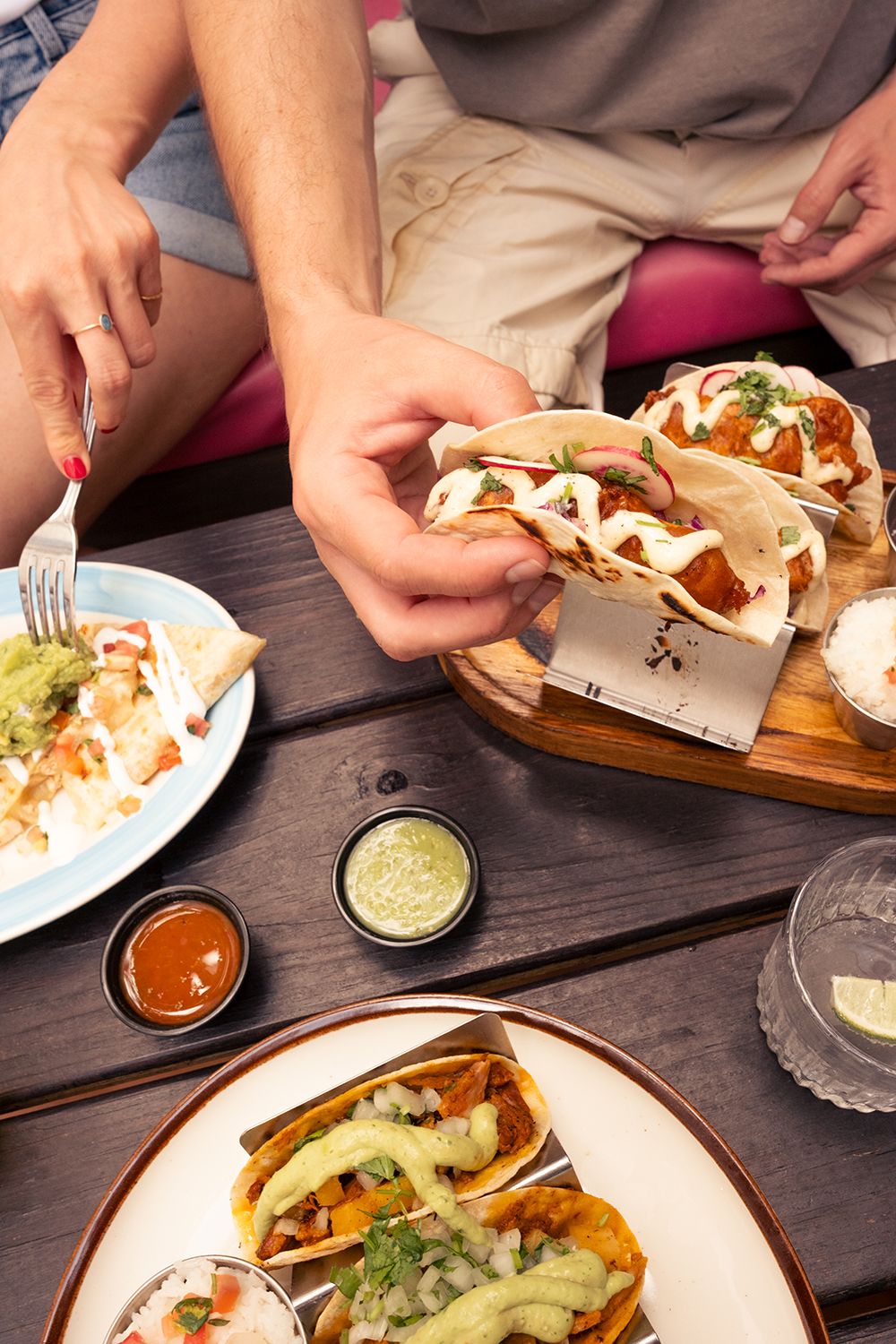 People enjoying tacos with toppings, nachos, and sauces on a wooden table, sharing a relaxed dining experience