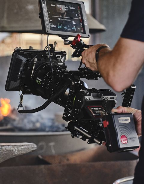 A person operating a professional video camera rig with an attached monitor, documenting a scene with flames in the background