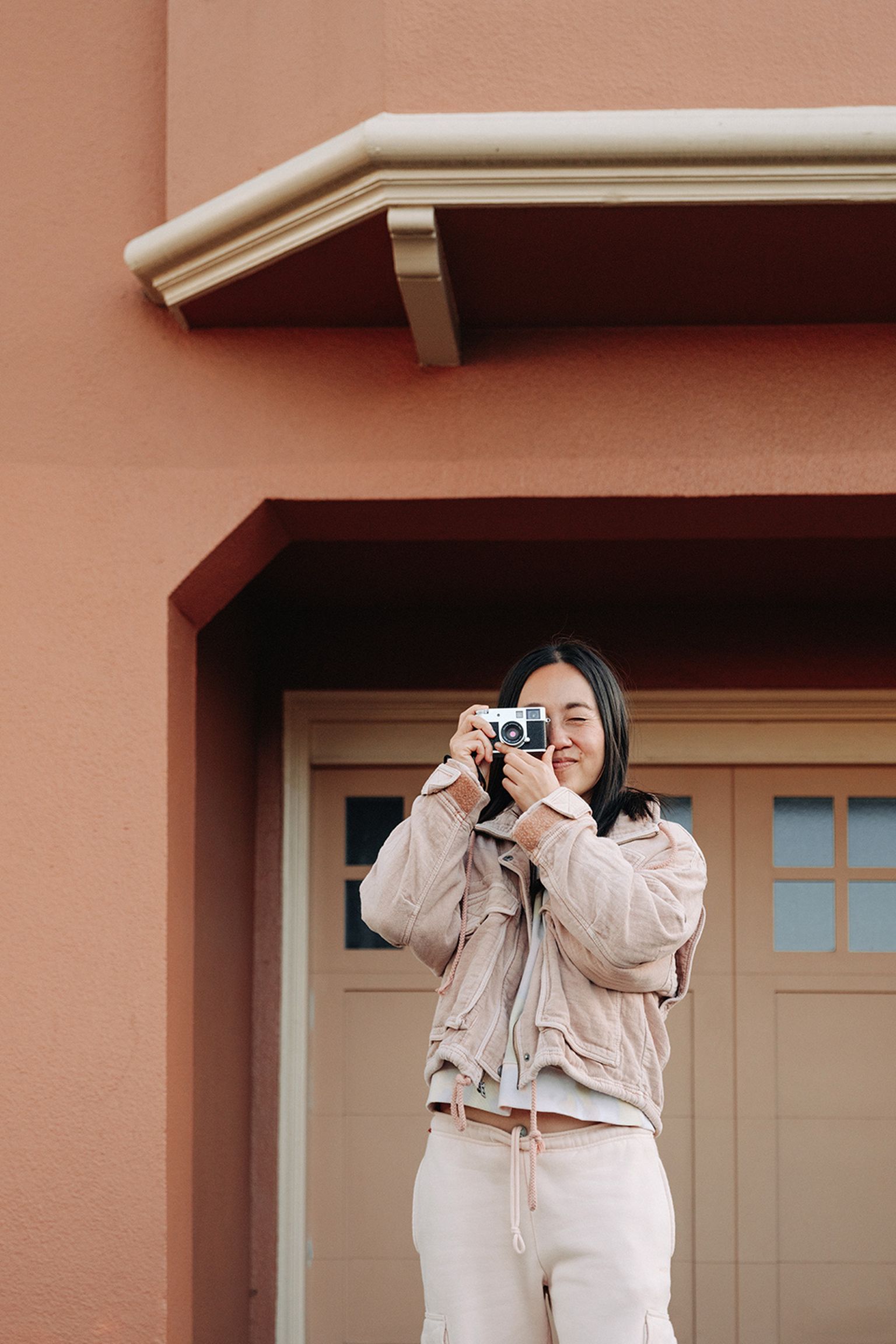 A person stands in front of a peach-colored building, holding a camera up to their eye