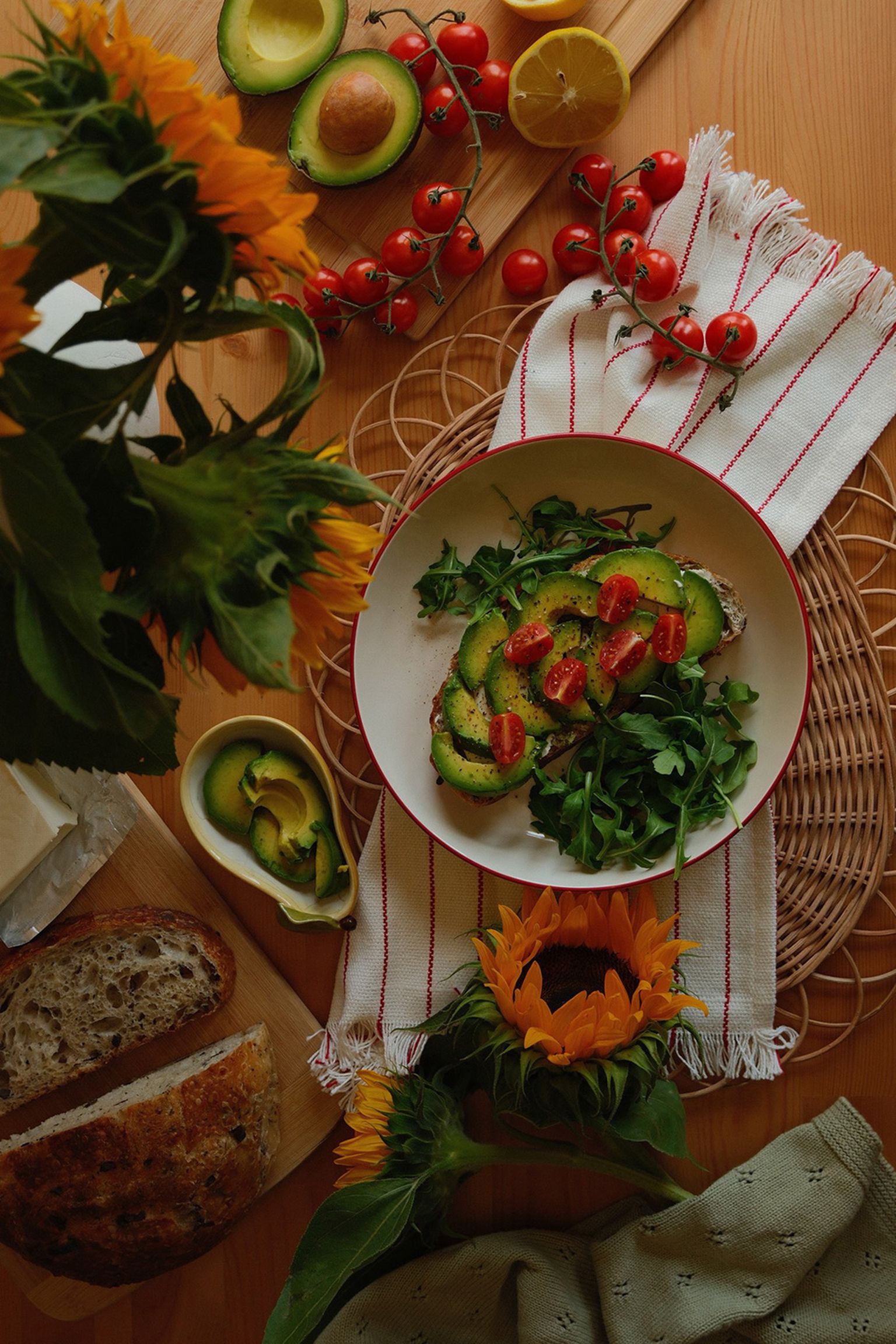 A table with avocado toast and arugula on a plate, surrounded by cherry tomatoes, sliced bread, sunflowers, and avocado halves on a wooden surface