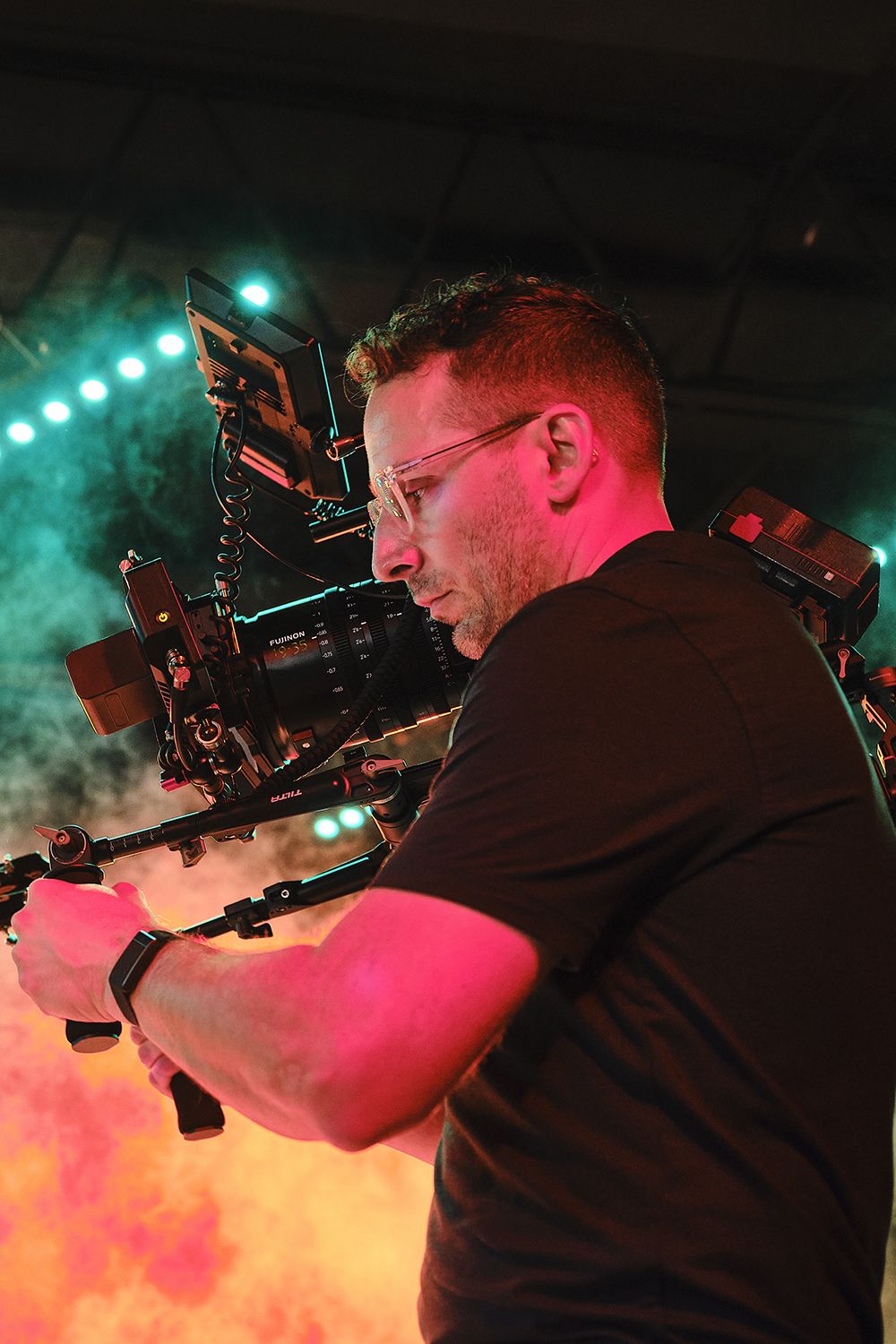 A man wearing glasses operates a large video camera, surrounded by colorful stage lighting and smoke effects