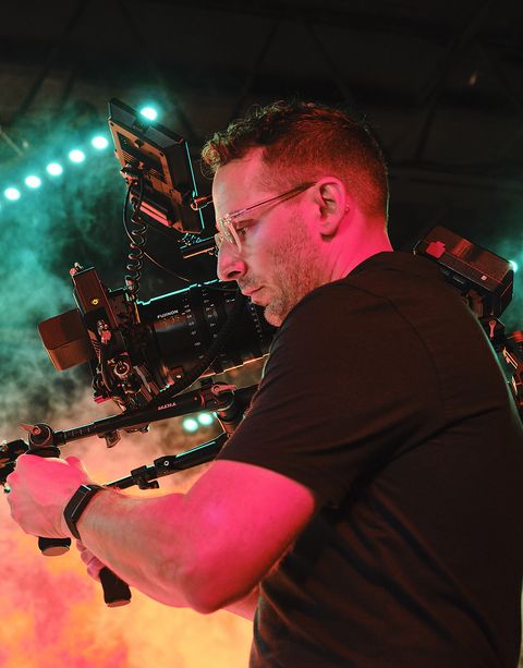 A man wearing glasses operates a large video camera, surrounded by colorful stage lighting and smoke effects