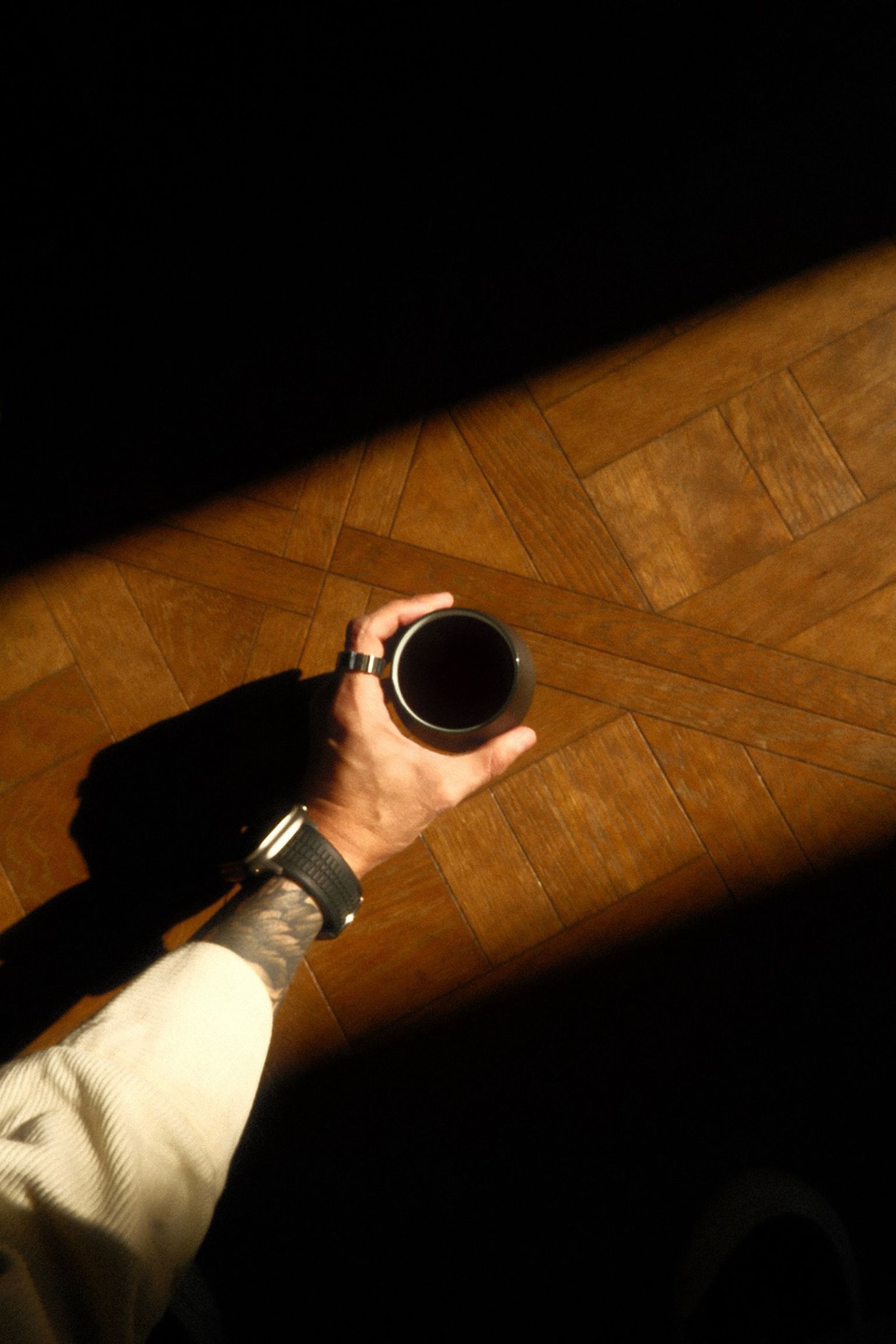 A person in a light shirt holding a cup of coffee, standing on a wooden floor with a pattern, partially illuminated by sunlight