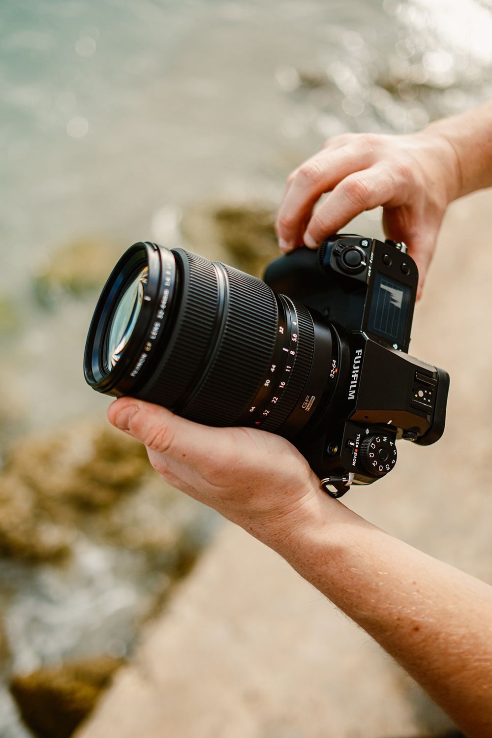 Hands holding a camera with a large lens by the water, focused on the camera's details and settings