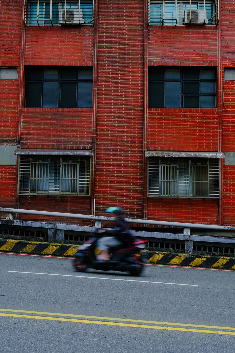 A person on a motorbike speeds past a red brick building with barred windows on a deserted road