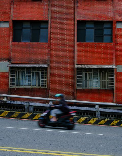 A person on a motorbike speeds past a red brick building with barred windows on a deserted road