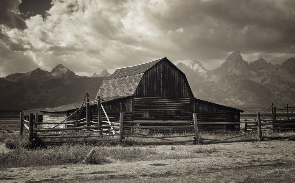 Sepia-toned image of a rustic barn with a wooden fence, set against a backdrop of dramatic clouds and distant mountains
