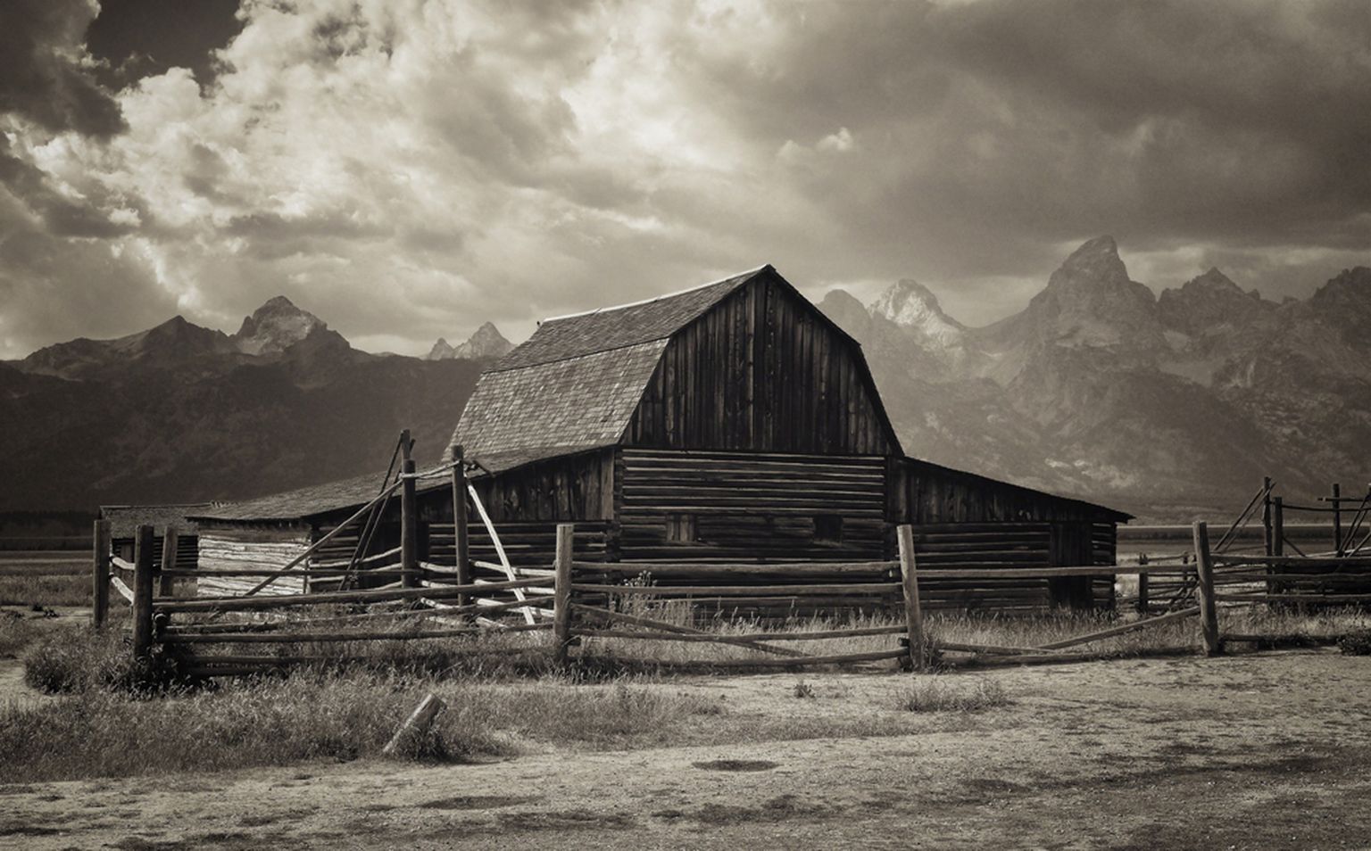 Sepia-toned image of a rustic barn with a wooden fence, set against a backdrop of dramatic clouds and distant mountains