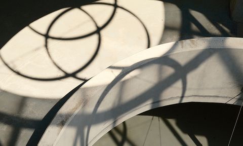 Shadows of circular metal structures cast intricate patterns on a curved concrete surface in sunlight.