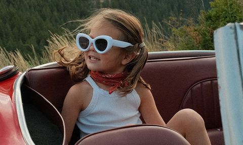 A young girl with sunglasses and a bandana sits in a red convertible, smiling, with a forested landscape in the background.