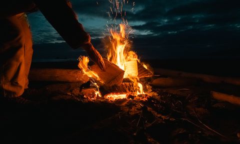 A person adds wood to a campfire on a beach at dusk, with sparks flying and a dark, cloudy sky in the background.