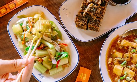 A person uses chopsticks to pick up vegetables from a dish. Nearby is a plate of fried tofu and a dish with sauce.