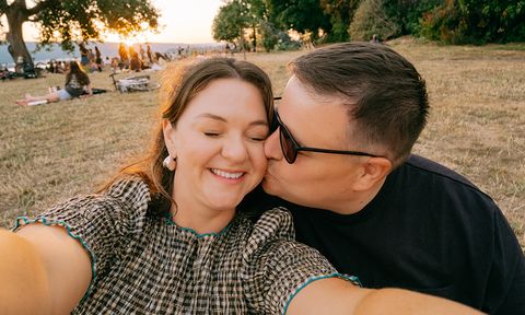 A couple creates a selfie outdoors at sunset; the man kisses the woman's cheek as she smiles. People and trees are in the background.