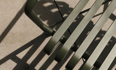 Close-up of a green metal chair casting striped shadows on a concrete surface in sunlight.