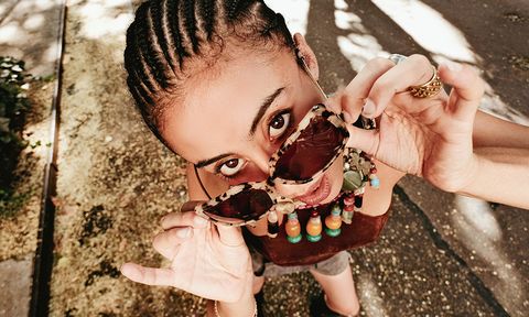 Person with braided hair playfully holding sunglasses, looking up. Wearing colorful beaded necklace and rings, standing on a sunlit street.
