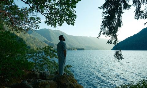 A person stands on a rocky lakeshore, gazing at the serene water and distant mountains under a clear sky, surrounded by lush trees.