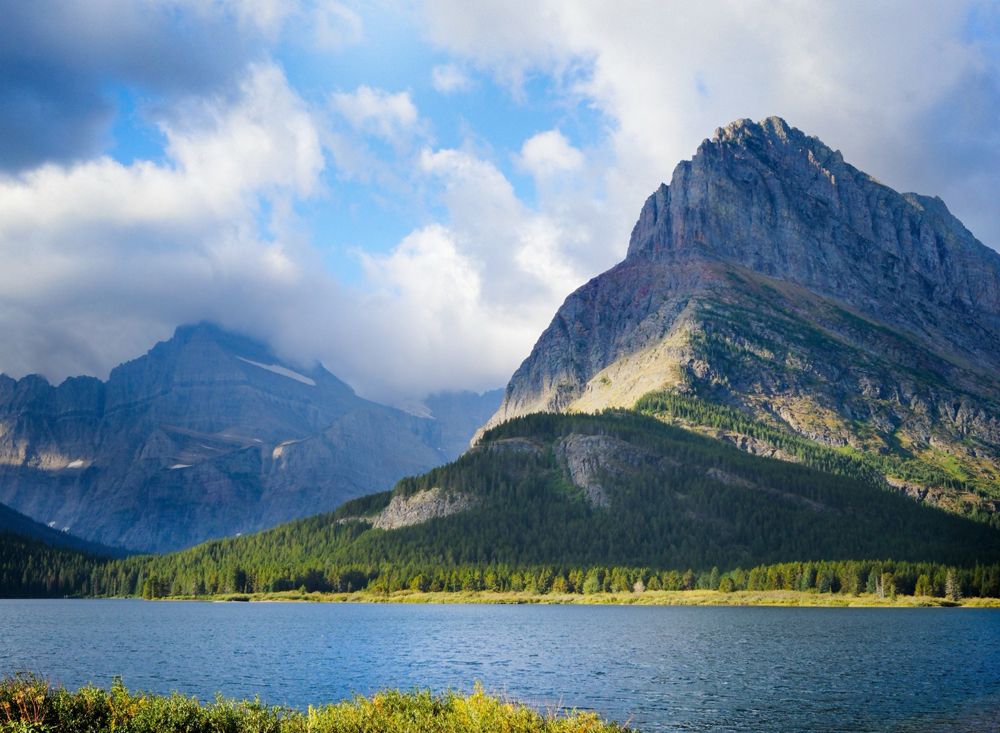 Mountain landscape with a clear lake in the foreground, lush green forests on the slopes, and sunlight illuminating the peaks under a partly cloudy sky