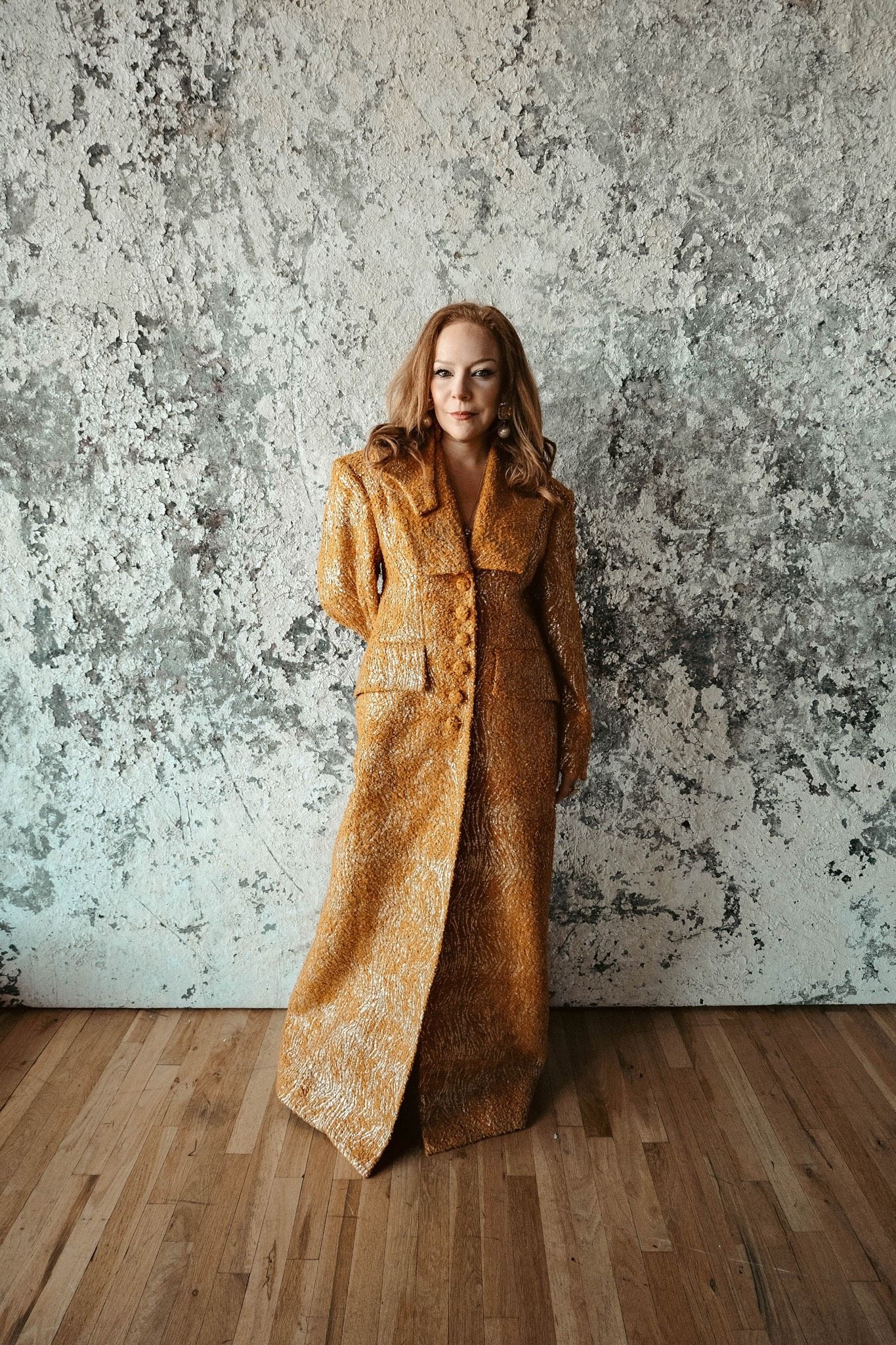 A person in a long, textured golden coat stands on a wooden floor against a distressed concrete wall