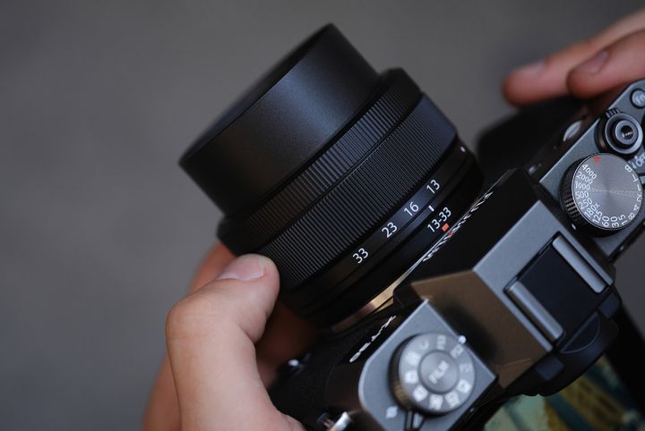 Close-up of hands holding FUJIFILM X-T30 III, focusing the lens. The lens has aperture settings visible, and the camera has dials and buttons on top.