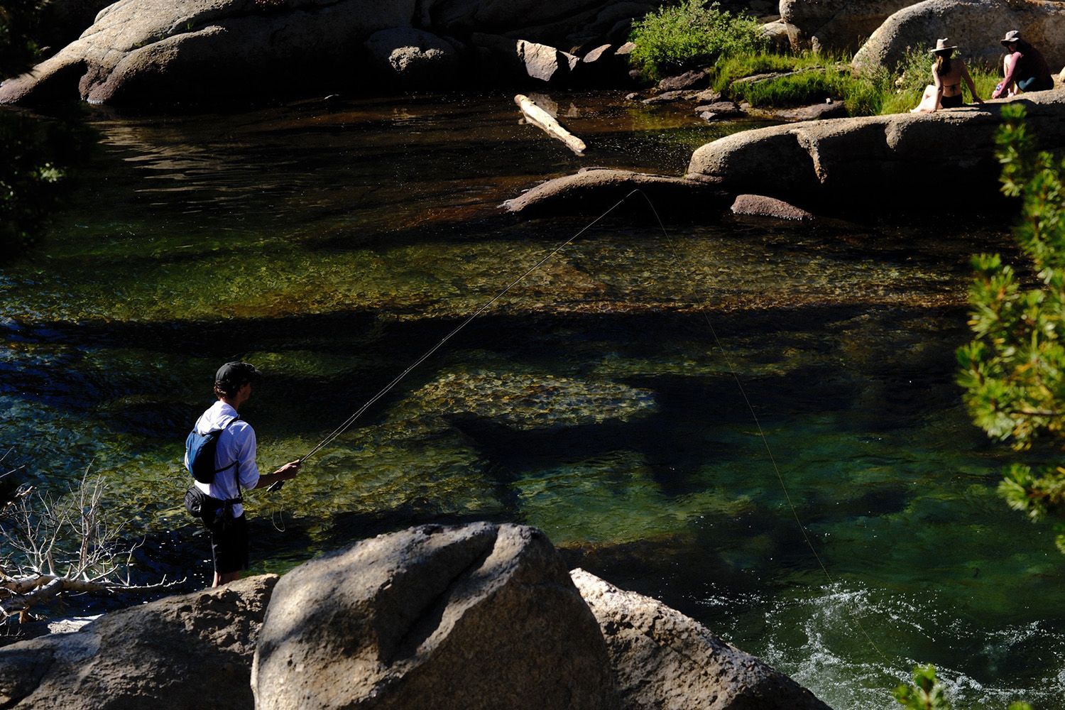 A person fishing in a clear, rocky river surrounded by lush greenery, with two people sitting on rocks in the background