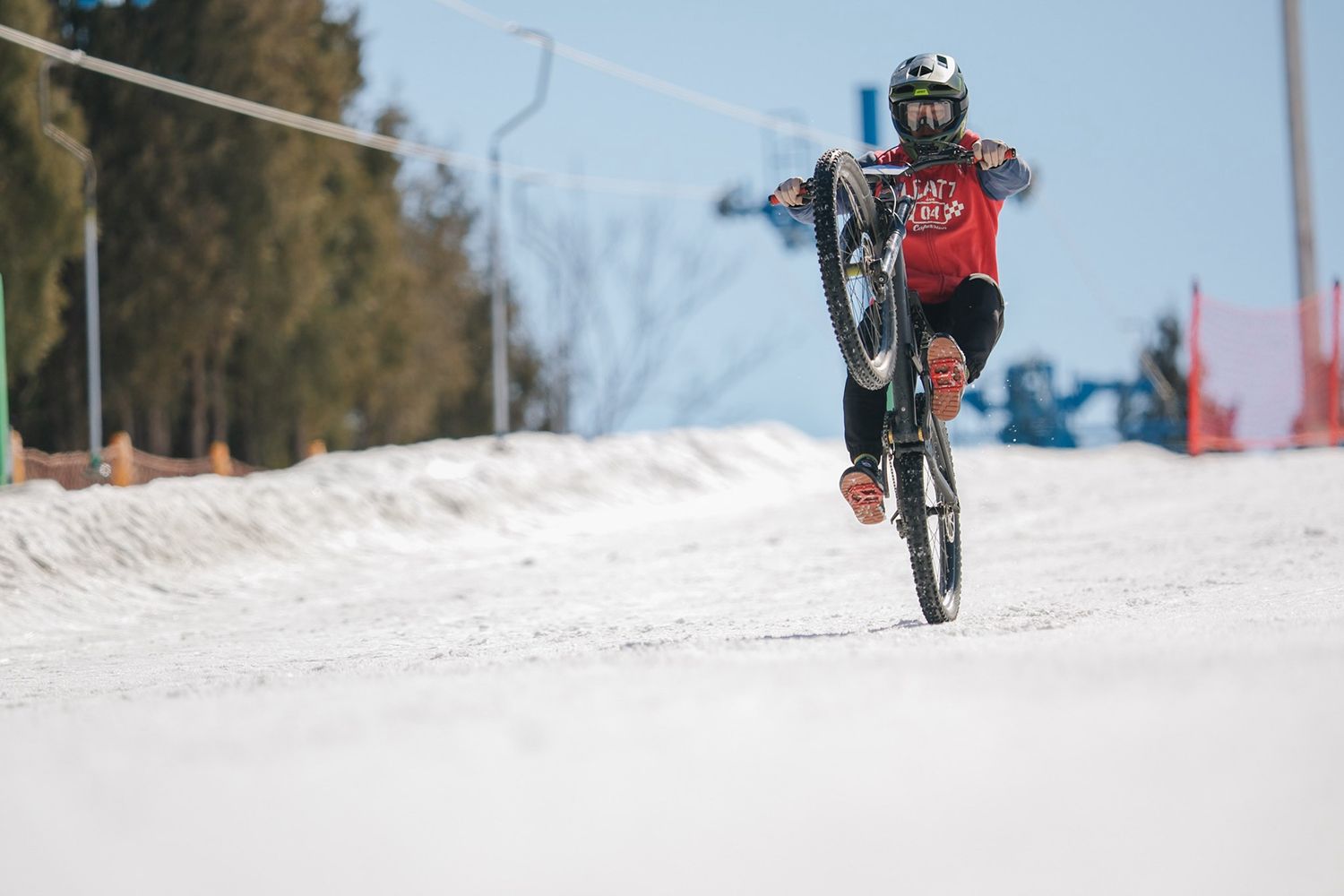 Person in a helmet and red shirt performing a wheelie on a bicycle on a snowy path, with trees and distant ski lifts in the background