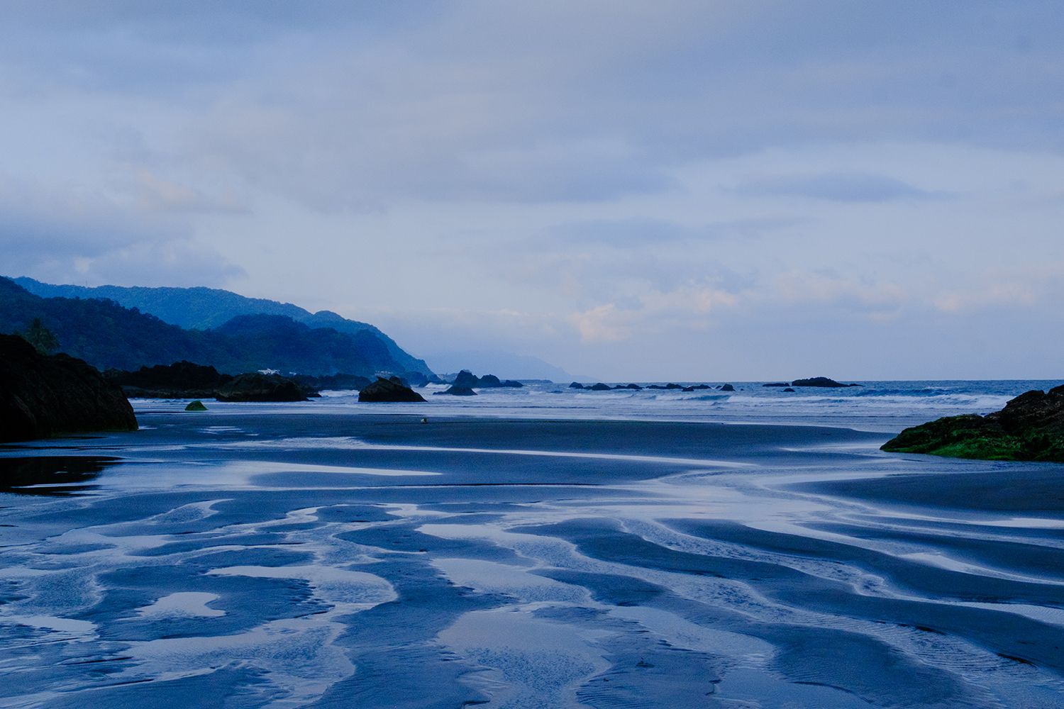 A serene coastal scene with rippled sand patterns, distant mountains, and a calm sea under a cloudy sky, all in a blue hue