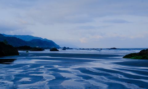 A serene coastal scene with rippled sand patterns, distant mountains, and a calm sea under a cloudy sky, all in a blue hue