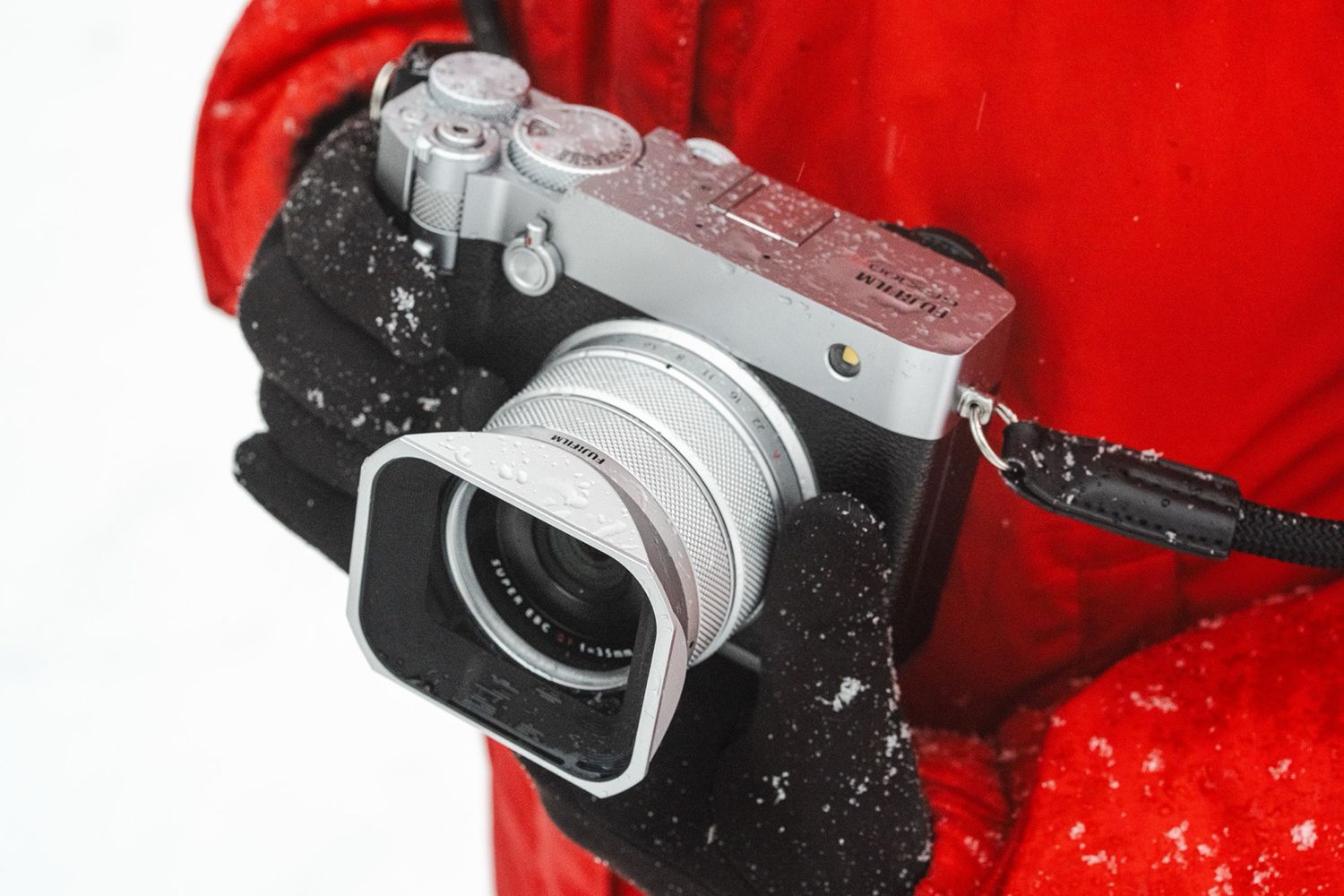 Person holding a camera with a square lens hood, wearing a red coat and black gloves dusted with snow