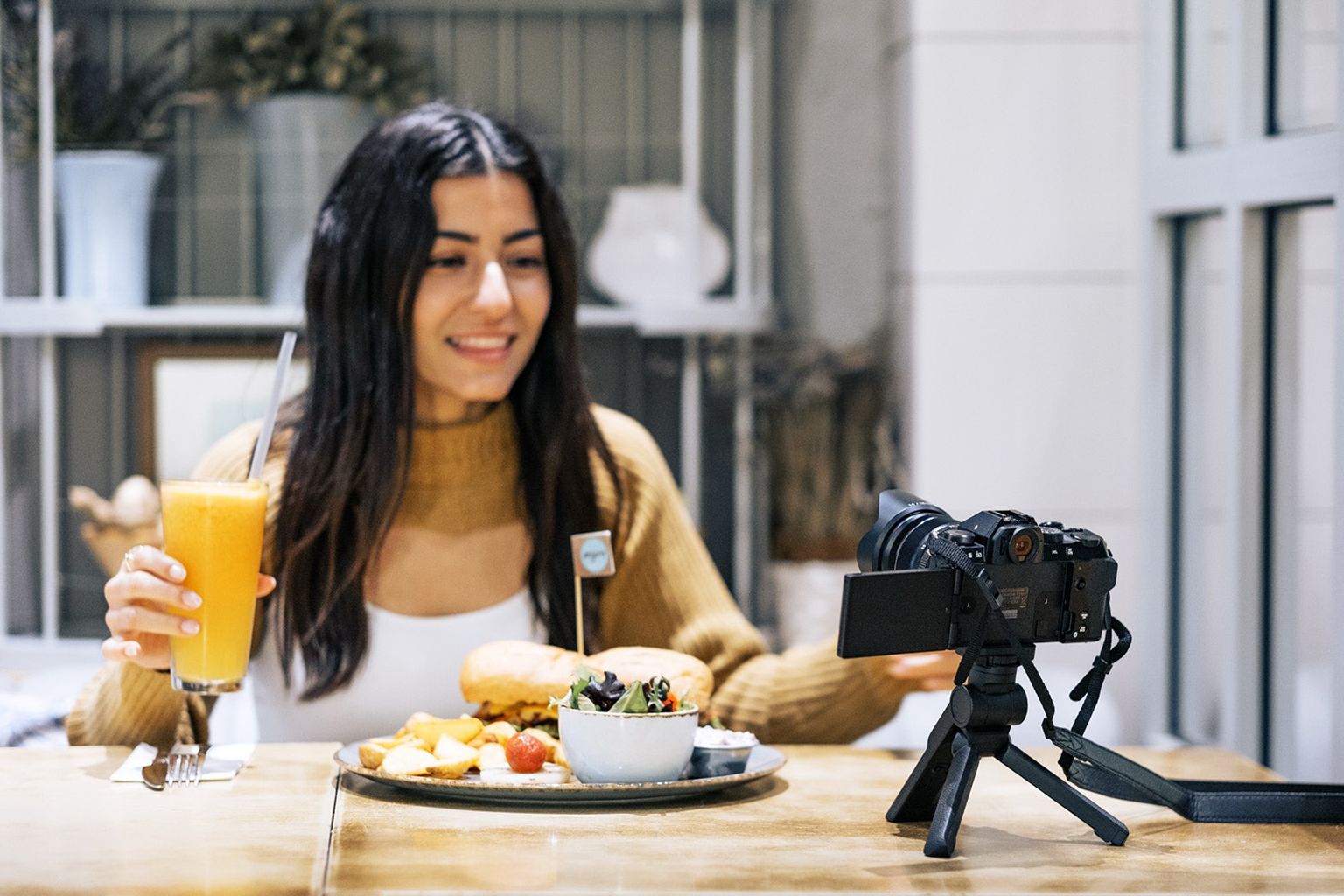 A woman sits at a table with a burger and orange juice, smiling at a camera on a tripod
