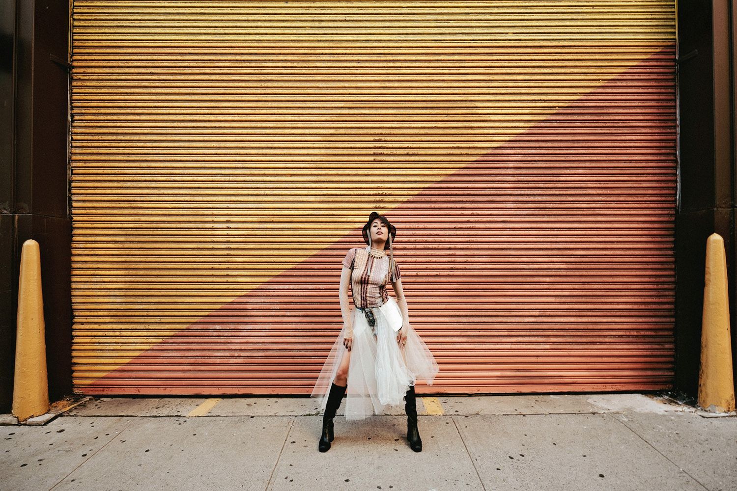 Person in eclectic fashion poses in front of a large yellow and pink diagonal-striped corrugated metal door