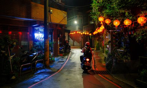 Narrow alley at night with a person on a scooter. Red lanterns illuminate the scene, creating a warm glow amid neon lights